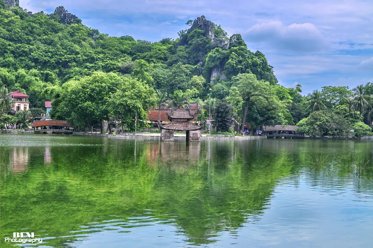 A small pavilion standing on a calm lake in front of green forested limestone hills and a cloudy blue sky