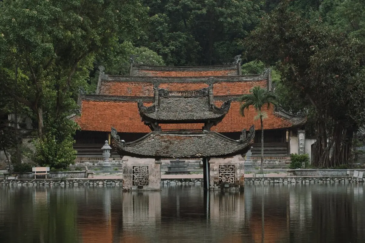 A small stone water pavilion with a curved grey tile roof standing in a lake, in front of the orange-tiled roofs of Thầy Pagoda and dark green trees